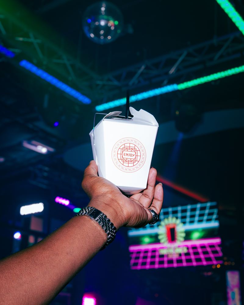 A Tokyo Joe's cocktail served in a Chinese takeout box, held up in front of the neon grid